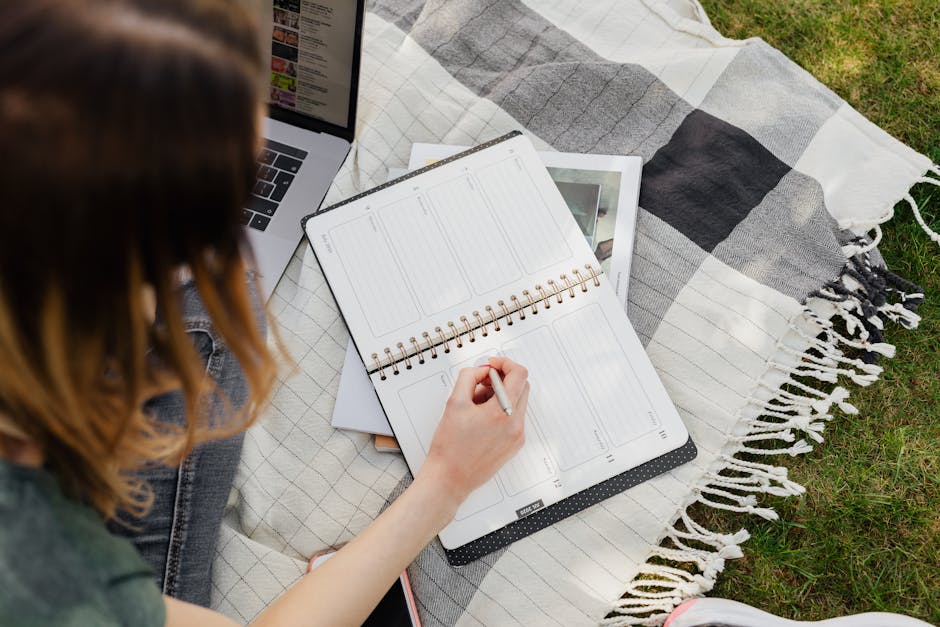 From above of crop female college student sitting on blanket placed on green lawn and writing down important information in planner with open laptop beside on fine summer day